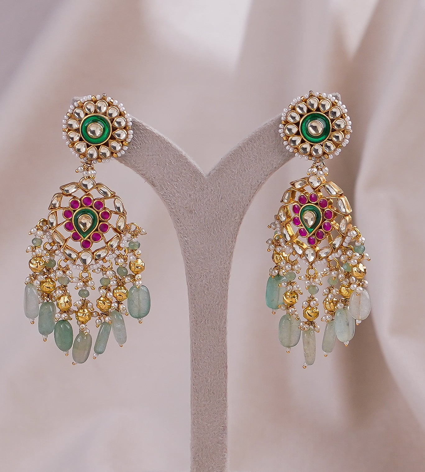 Gold jhumka earrings with green and pink stones, pearls, and dangling beads on display stand