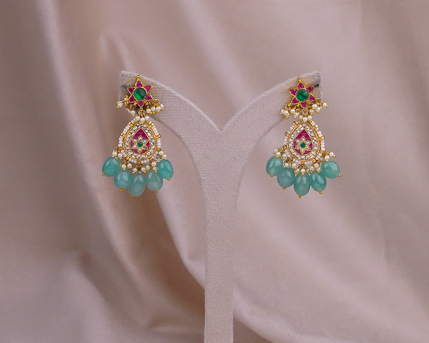 Gold jhumka earrings with emerald, ruby, and pearl detailing, displayed on a velvet stand