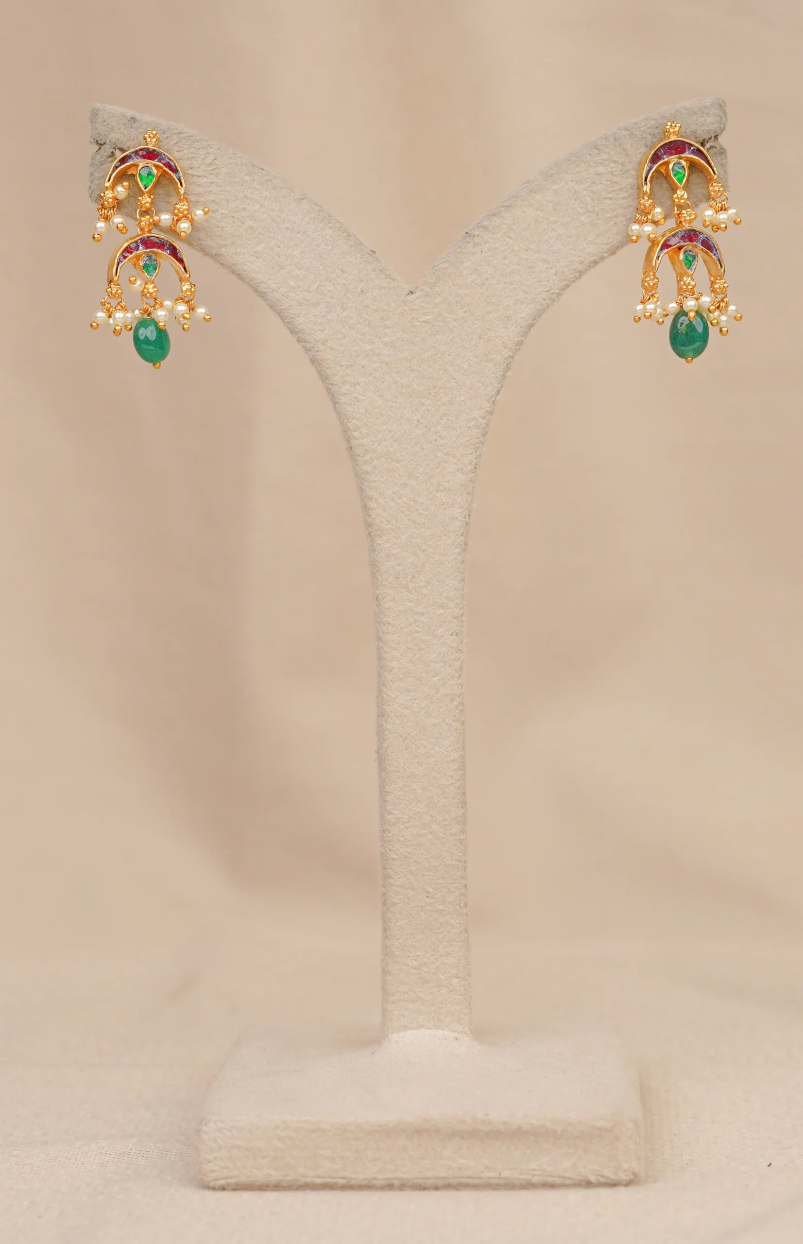 Gold jhumka earrings with green gemstones, red enamel, and pearl accents on a display stand